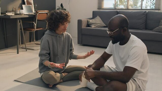 Full shot of Caucasian woman with curly hair sitting on mat and talking with African American man wearing glasses while resting and having water break after home workout