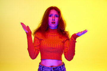 Young girl standing with unhappy face, spreading hands with shock against yellow studio background...