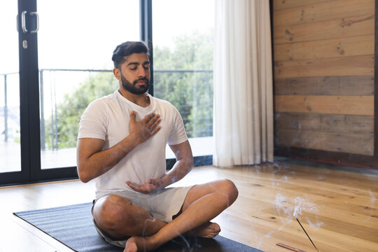 Relaxed biracial man sitting, practicing yoga, closing eyes and meditating in sunny room at home - Powered by Adobe