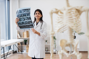 Confident indian doctor holding MRI scans of human brain in consulting room with skeleton model on blurred foreground. Professional female physician in medical uniform analyzing diagnostic report.