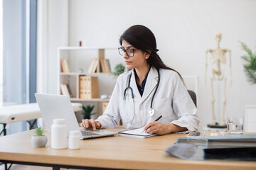 Concentrated female doctor with stethoscope sitting at table and writing prescription while using...