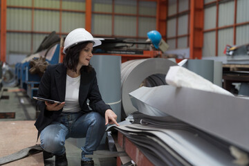 Female factory worker working using digital tablet inspecting quality of rolls of galvanized or metal sheet in aluminum material warehouse. Female inspector checking product material in factory - obrazy, fototapety, plakaty