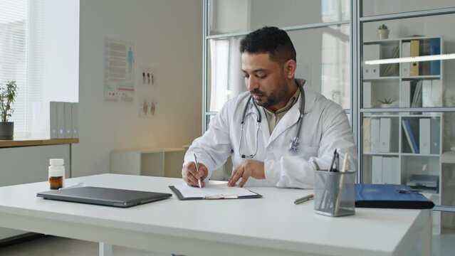 Waist up of Middle Eastern male practitioner wearing lab coat and stethoscope sitting at desk in his office and doing medical paperwork