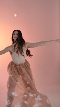 A young woman, a ballerina girl dancing, poses in a photo studio at a photo shoot, gently moving her arms and fingers on a pink background, slowmo, medium. Lady brown-haired woman with long hair