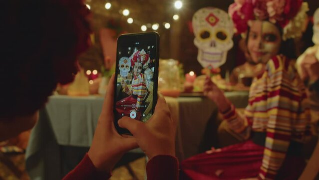 Over the shoulder of woman taking photos on smartphone of little girl with half painted face and flower wreath posing with paper skull mask, sitting at table in backyard on Dia de Muertos