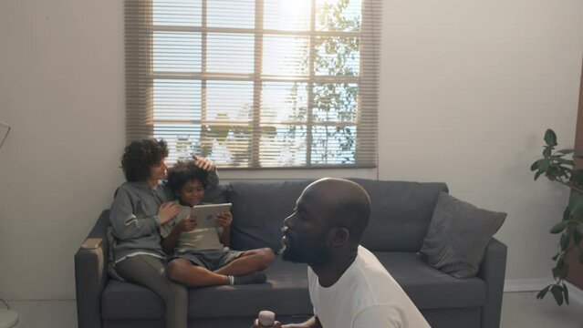 Medium shot of interracial family of mother and son sitting on couch watching movie on digital tablet while father doing squats with dumbbells