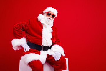Photo of good mood funky grandfather in santa costume in glasses sit on white platform hand on waist isolated on red color background