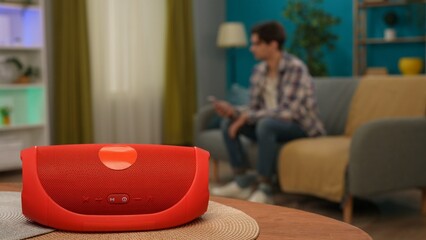 Man sitting on the sofa in the living room holding smartphone, red music speaker in focus on the table. Smart home concept.