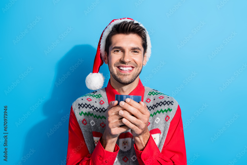 Sticker Portrait of toothy beaming person dressed santa hat hold cup of aromatic coffee in christmass morning isolated on blue color background