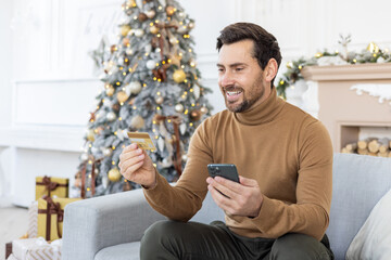 A smiling young man is sitting on the sofa at home, holding a mobile phone and looking at a credit card
