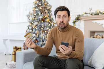 Credit card and billing issues. Portrait of a worried young man sitting at home on New Year's holidays, holding a phone and a credit card, spreading his hands and looking sadly at the camera