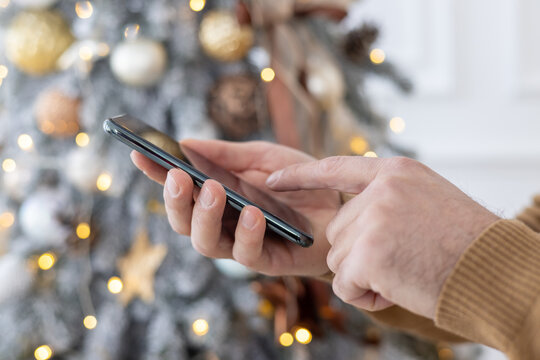 Close-up photo of young male hands in a brown sweater using a mobile phone against the background of a Christmas tree