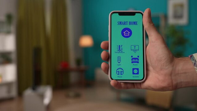 Man switching lamp, air conditioner, robot cleaner, opens curtains with smartphone app, focus on screen interface icons. Smart house concept.