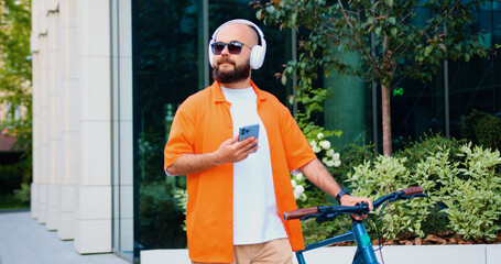 Portrait caucasian man going near business centre carrying bicycle using smartphone. Portrait of confident young male in stylish casual clothes with modern bike on city street. - Powered by Adobe