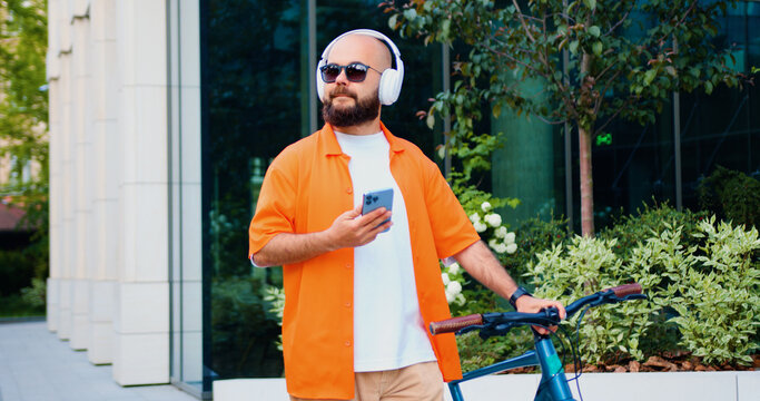 Portrait caucasian man going near business centre carrying bicycle using smartphone. Portrait of confident young male in stylish casual clothes with modern bike on city street.