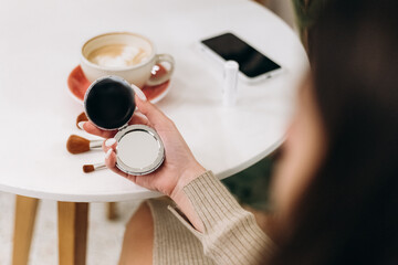 Young pretty woman sitting inside cafe using brushes and mirror for daily makeup, relaxing smiling happily, joyful posing enjoying atmosphere near plant flowerpot

