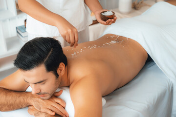 Blissful man customer having exfoliation treatment in luxury spa salon with warmth candle light ambient. Salt scrub beauty treatment in health spa body scrub. Quiescent