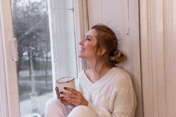 Close-up portraits of young stylish woman sitting on windowsill, drinking cocoa from cup, looking out window. Winter festive atmosphere. Girl in home clothes drinks tea, coffee. Holiday light bulbs.