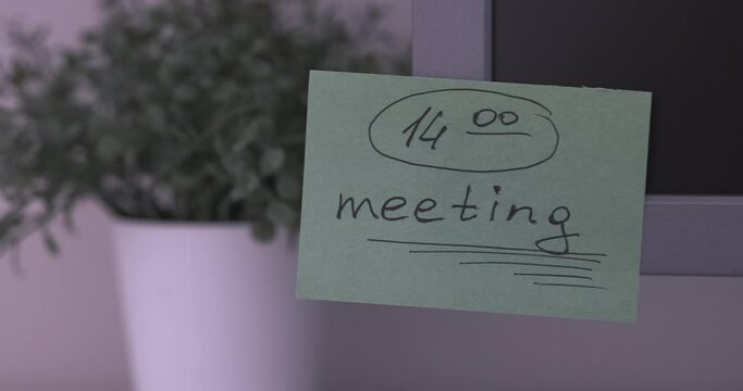 A green paper notes with the reminder 14-00 Meeting on it sticked on to a monitor at an office workplace.