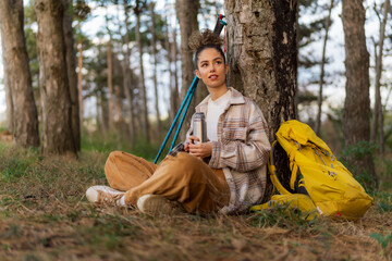 On the trail, a caucasian girl with curly hair rests against a tree, checking her phone as her backpack stands close.