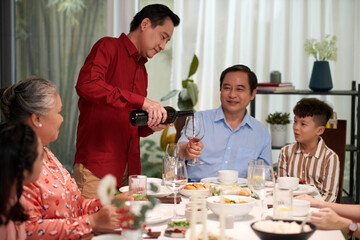 Smiling Vietnamese man pouring wine in glass of brother at family dinner