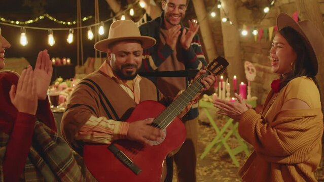 Medium shot of Mexican man playing guitar while having party on Day of the Dead with family members and friends, dancing and singing together in backyard decorated with candles and marigolds
