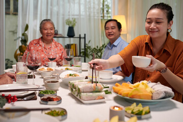 Smiling woman taking spring roll and bowl of sauce at family dinner - obrazy, fototapety, plakaty