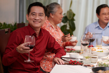 Cheerful mature Vietnamese man drinking glass of wine at dinner table with his family
