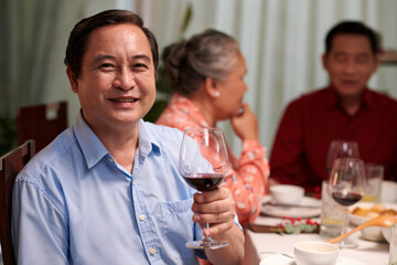 Portrait of cheerful Asian man toasting with glass of wine when sitting at dinner table with family members