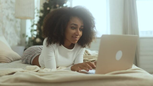 Charming young woman typing on computer order online shopping sitting near decorated tree indoors Pretty female working or studying from home on a laptop during Christmas holidays sitting on bed alone
