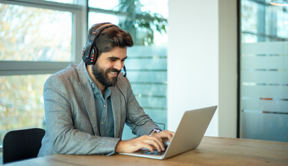 Young middle eastern businessman working on laptop computer wearing headphone at modern office. Smiling business man wearing headset for video conference, communication, education and e-learning.