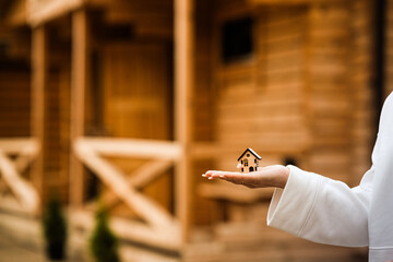 A small wooden house on a woman s hand against the background of a built wooden house. Concept of buying an ecological house made of wood