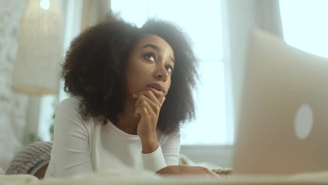 Portrait of pretty creative african american woman student thinks questioningly about difficult choice choose imagine plan in mind has doubts create idea at desk laying on bed at home