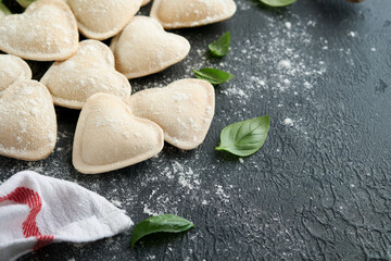 Fototapeta premium Italian ravioli pasta in heart shape. Tasty raw ravioli with flour and basil on dark background. Food cooking ingredients background. Valentines or Mothers Day lunch ideas. Top view with copy space.