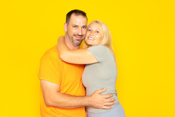 Real Caucasian couple in their 40s tenderly hugging each other demonstrating their love and commitment, isolated on yellow background.