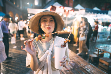 Fototapeta premium Happy young asian foodie woman eating bbq grilled skewers at outdoor night market street food vendor