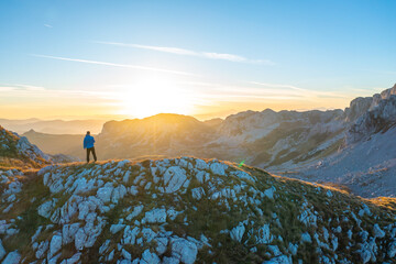 A hiker standing on a hill and looking at the beautiful sunrise behind the mountain peaks in the distance.