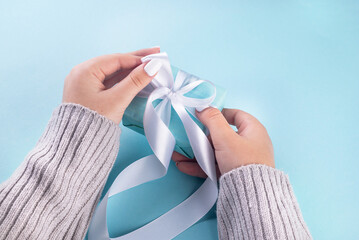 First person top view photo of female hands holding blue gift box with white ribbon. A pastel blue setting welcomes your words. my hands cradle a blue gift box