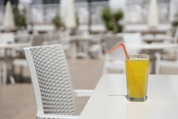 Glass of orange juice with plastic straw on white table in restaurant outdoor lounge zone in sunny weather