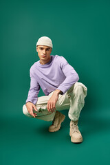 full length of man in purple sweatshirt and beanie hat sitting on turquoise backdrop, winter fashion