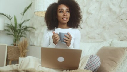 Pretty young curly woman freelancer with cup of hot drink working online typing browsing on laptop sitting on bed indoors Positive female enjoying great day at cozy bedroom at home
