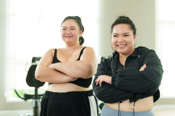 Fototapeta na wymiar Two chubby Asian women wearing sports clothes stand smiling happily in the gym, both arms crossed and looking at each other. Women's happiness after exercise. Concept Lifestyle and exercise