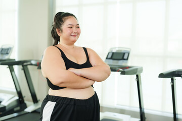 Fototapeta premium Beautiful overweight Asian woman stands with arms crossed and proudly smiling as gives a thumbs up to the camera and gives a thumbs up standing in village gym. Lifestyle and exercise concepts