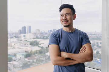 Fototapeta premium Handsome Asian man with beard smiling and standing at apartment room alone with city view, successful happy living life, look outside.