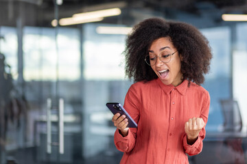 Young successful businesswoman boss received online notification on phone victory, good results of achievement, woman celebrates victory and triumph, happy holds smartphone in hands, uses app.