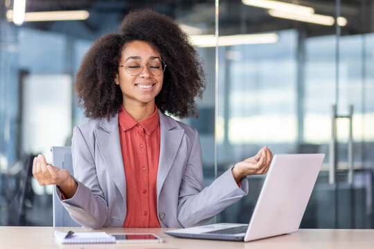 Young joyful businesswoman meditating indoors lotus pose, boss business suit smiling satisfied with closed eyes inside office sitting laptop, african american woman achievement results, dreaming.