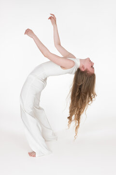 Full length shot barefoot young woman with eyes closed having fun while bending over backwards on white background. Side view of flexible blondie female model with long wavy hair dressed in jumpsuit