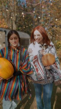 In the autumn garden, two pretty girls in hippie costumes are carrying pumpkins.