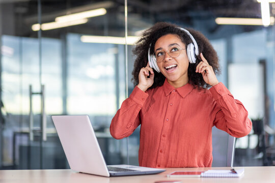 Young successful joyful woman working inside office with laptop, female programmer listening to music, singing along and dancing while sitting at workplace. Business woman in a shirt with curly hair.