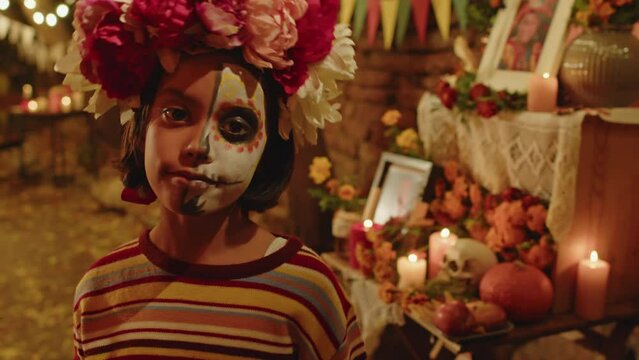 Medium closeup portrait of little Hispanic girl with half painted face and flower wreath on her head posing with craft skeleton mask in backyard at night during Day of the Dead celebration
